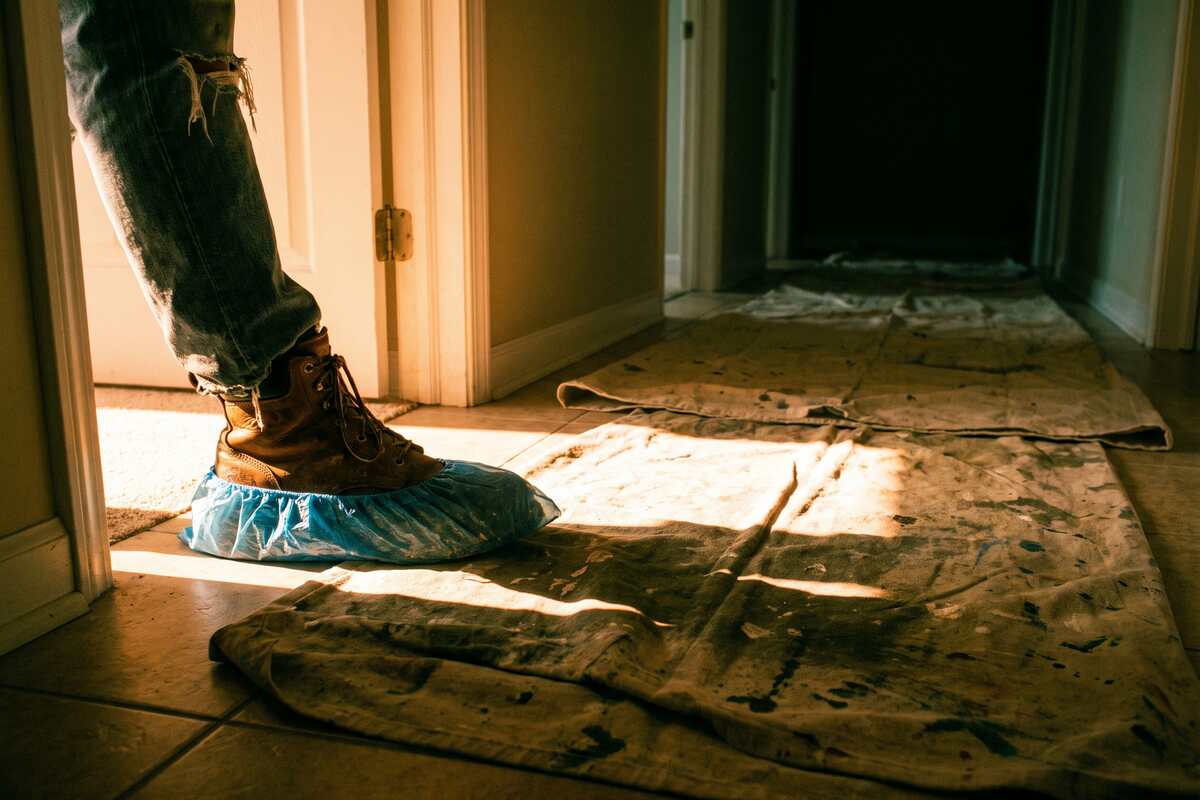 Blue shoe covers and a canvas drop cloth laid down on a tile hallway floor next to a toolbag.